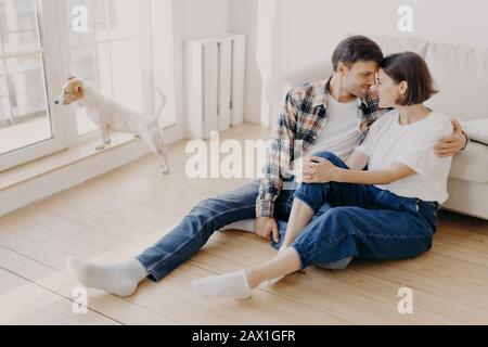 Horizontal shot of pleased husband and wife embrace and kiss, have good relationship, sit on wooden floor near couch, enjoy being at home during weeke Stock Photo