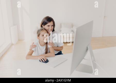 Indoor shot of happy lovely mother and daughter watch funny cartoon on computer, have pleasant smiles, enjoy togetherness, spend free time or weekend Stock Photo