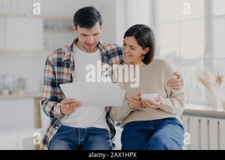 Caring man embraces his wife, look through documents with happy expressions, study payment bills, drinks coffee, dressed in casual wear, pose in spaci Stock Photo