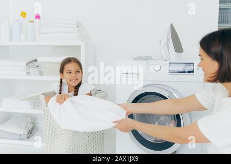 Happy girl with two pigtails poses in basket with dirty linen, has fun in laundry room with mother, helps to do washing. Woman loads washing machine, Stock Photo