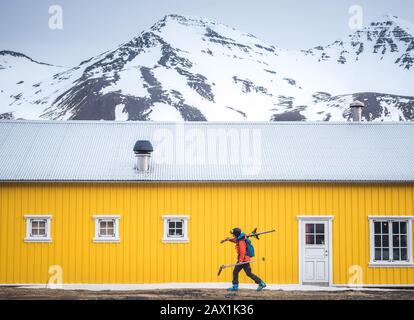 A woman walking with skis in front of a yellow building with mountains Stock Photo