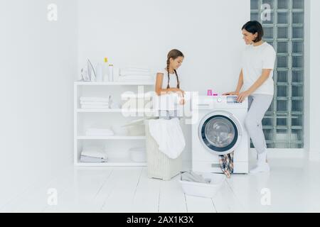 Indoor shot of happy mother and daughter stand near washing machine, girl pours liquid powder, load washer with dirty clothes, do housework, have laun Stock Photo