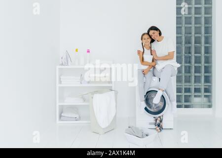 Shot of beautiful woman and his small daughter embrace and smile pleasantly, sit on washing machine, wash linen in laundry room, have friendly relatio Stock Photo