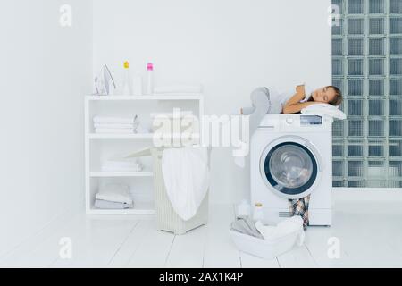 Female preschooler sleeps on washing machine, being tired with washing, poses in white big laundry room with basket and basin full of dirty clothes bo Stock Photo