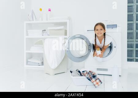 Small girl with appealing appearance, has fun and poses inside of washer, holds detergent, prepares for washing, basin with clothes for putting into w Stock Photo