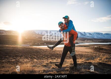Man carrying woman on piggyback in field with mountains in background Stock Photo