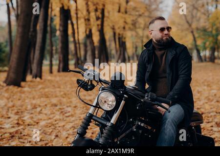 Outdoor shot of pensive male biker keeps gaze aside, has thoughtful look, wears sunglasses, sweater, black jacket and jeans, poses on motorbike outdoo Stock Photo