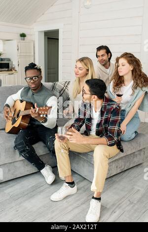 Vertical shot of friends sitting on sofa in modern apartment listening to guy playing acoustic guitar music Stock Photo