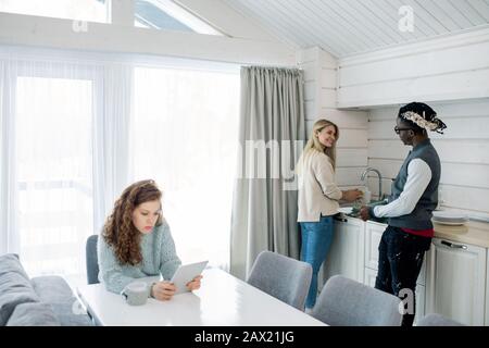 Blond woman chatting with Black guy while washing dishes, their friend sitting at table and reading something using tablet PC Stock Photo