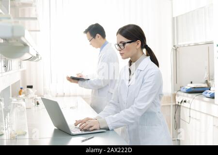 Horizontal portrait of two young laboratory scientists wearing white coats making notes in papers and on laptop Stock Photo