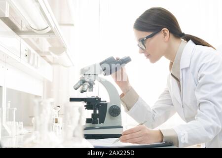 Horizontal side view shot of young female medical scientist examining specimen using microscope in modern laboratory Stock Photo