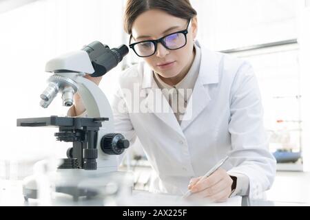 Young female medical worker making notes after examining specimen using microscope in modern laboratory Stock Photo