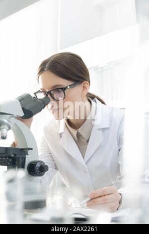 Vertical portrait young Caucasian medical scientist wearing eyeglasses examining specimen using microscope in modern laboratory Stock Photo