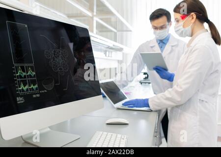 Horizontal shot of two scientists wearing white coats, masks and gloves working in laboratory using computers and gadgets Stock Photo
