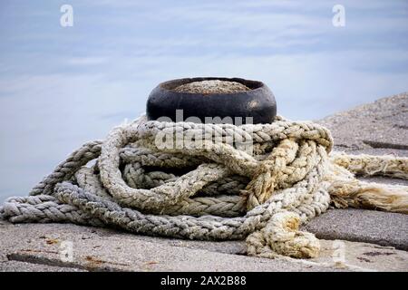 Mooring metal bollards with old, damaged knit rope tide around it on the stone marine dock in the cloudy day Stock Photo