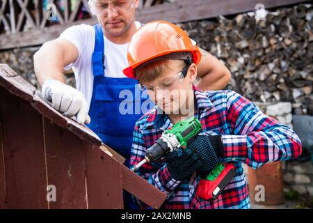 Cute boy in a construction helmet and glasses with a screwdriver in his hands building wooden dog house with his father. Construction and decoration o Stock Photo