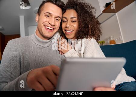 Romantic Date. Young multiethnic couple at home sitting on sofa using digital tablet together laughing joyful close-up Stock Photo