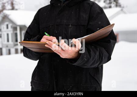 an unrecognizable man standing outside during home inspection on snow falls. he wearing a cold winter black jacket as he taking notes on notebook Stock Photo