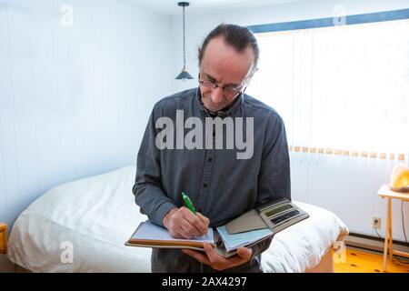 middle aged home Inspector taking notes during an inspection. A man with glasses writes on a notebook, with the bedroom interior background Stock Photo