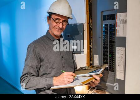 home Inspector in front of electric distribution board during an inspection, a caucasian man wearing helmet and glasses taking notes on his notebook Stock Photo