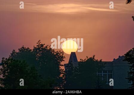 The sun on an early foggy morning over the roofs of houses. Orange sky. Stock Photo