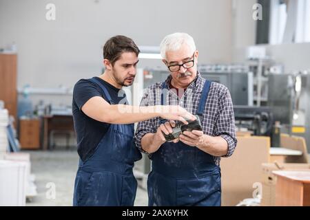 Young specialist in overall showing senior coworker how to use controller at factory Stock Photo