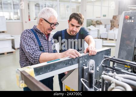 Senior worker in glasses explaining printing machine setups to young colleague at modern factory Stock Photo