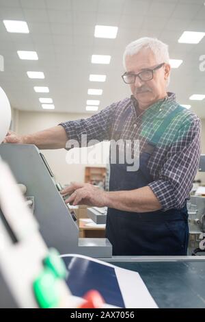 Serious senior printing worker with mustache pushing buttons on control panel while operating printing machine at factory Stock Photo