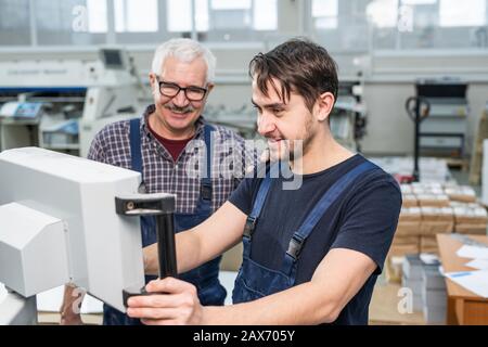 Smiling young bearded intern of printing factory operating machine under control of experienced specialist in workshop Stock Photo