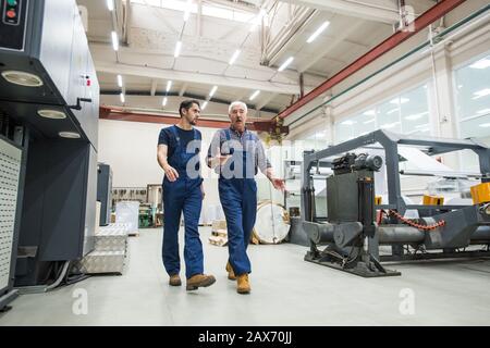 Experienced senior man in blue overall gesturing hands while interacting with young worker at printing factory Stock Photo