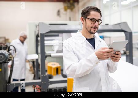 Content young bearded specialist in glasses using digital tablet while analyzing printing resources at plant Stock Photo