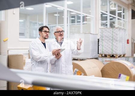 Senior and young lab specialists in white coats using digital tablet at factory while talking about industrial productivity Stock Photo