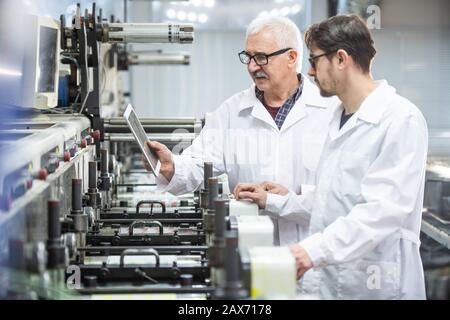 Serious senior and young quality inspectors in lab coats using tablet for analysis of printing press errors at printing factory Stock Photo