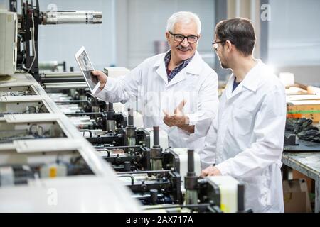 Positive senior inspector in lab coat talking to colleague while using tablet to analyze print quality Stock Photo