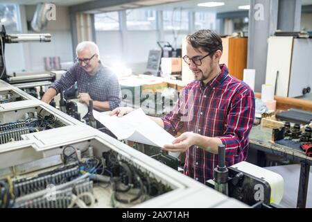 Smiling young worker in casual shirt standing at printing machine and checking printed paper Stock Photo