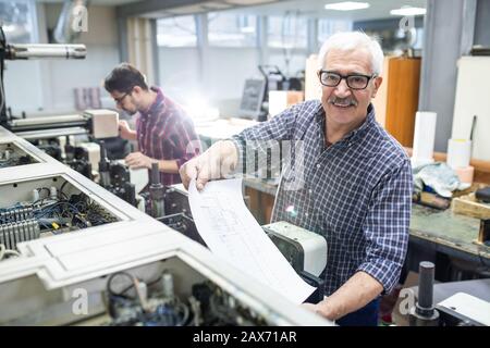 Portrait of cheerful senior man in glasses getting printed paper out of printing press at factory Stock Photo