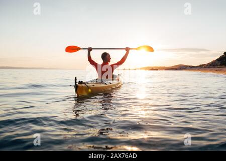 Rear view of man holds kayak paddles high. Active senior in a kayak on the sunset sea Stock Photo