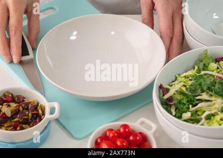 Woman preparing vegetable salad. Close up of fresh organic vegetables, cabbage mix, tomatoes, dried cranberries, and pumpkin seeds on a kitchen table Stock Photo