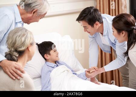 five year old asian child lying in bed in hospital ward gets a visit from parents and grandparents Stock Photo