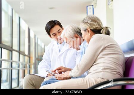 young asian doctor discussing test result and diagnosis with senior couple patients using digital tablet in hospital hallway Stock Photo