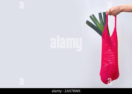hand holds red string bag with leek. Gray background with copy space. The trend concept is no plastic, recycling, zero waste, no cellophane. Stock Photo