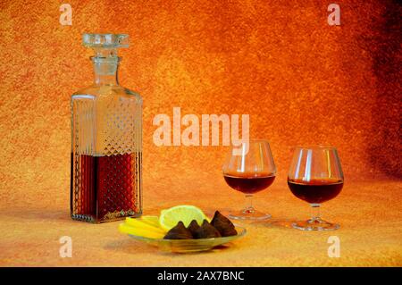 Crystal decanter with cognac and two glasses of drink next to a plate with appetizers, cut lemon and chocolate candies. Close-up. Stock Photo