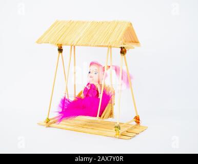Side view of a miniature handmade crafted doll in a pink dress on a swing made with toothpicks in-front a white background Stock Photo