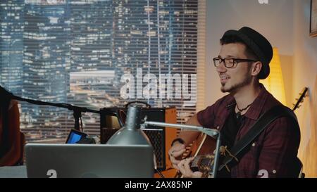 Profile view of handsome hipster singing into microphone and playing the guitar in home recording studio. There are skyscrapers in the background. Stock Photo