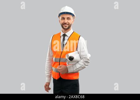 Front view of young architect in orange reflective vest and helmet holding big paper rolls. Smiling engineer carrying construction plans, isolated on grey. Concept of architecture, construction. Stock Photo