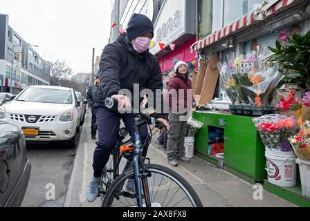 A Chinese American man wearing a surgical mask rides his bike on the sidewalk in Chinatown, Flushing, New York City. Stock Photo