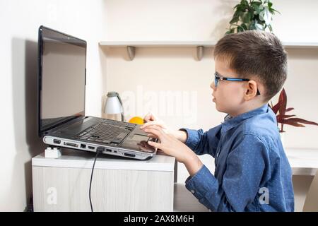 A boy in glasses and a blue shirt is sitting at a desk in front of a laptop and playing a computer game. Stock Photo