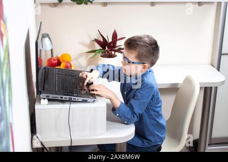 The boy is sitting at a desk in front of a laptop. Both hands on the keyboard touch the cursor control plate Stock Photo