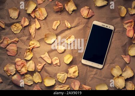 White smartphone with pink flower petals on crumpled kraft paper. Flat lay. Copy space on the phone. Suitable for layout. Vignetting. Stock Photo