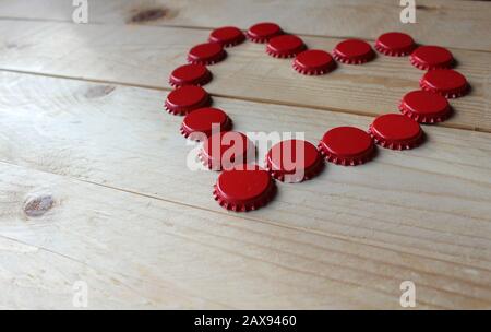 A red love heart made from beer bottle tops viewed from the side on a rustic wooden table. Beer drinkers Valentine's day concept. Selective focus. Stock Photo